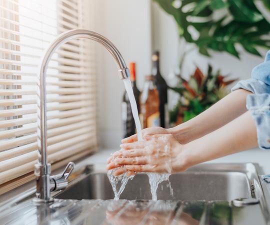 Woman washing hands in kitchen sink. The tap is on and you can see the woman's arms and hands. She is wearing a blue shirt.