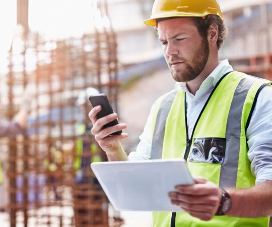 Man wearing a hi-vis vest and a yellow hardhat. He is looking at his mobile phone and also holding a tablet. 
